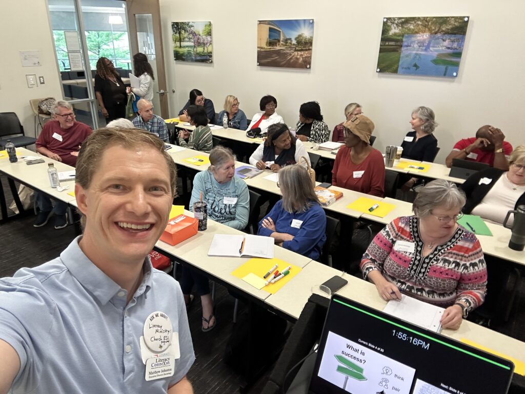 Literacy ConneXus Executive Director Matthew Johnston takes a selfie with participants during an ESL teacher training workshop, where volunteers are gathered around classroom tables for discussion and activities.